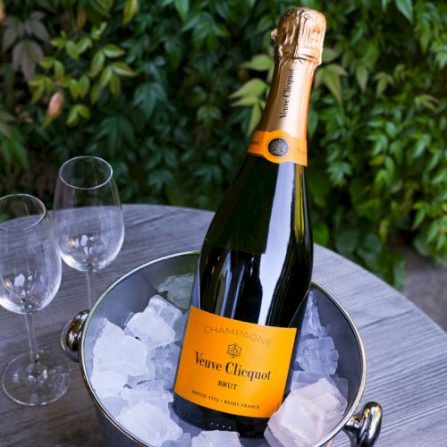A chilled bottle of Veuve Clicquot champagne in an ice bucket with two empty wine glasses on a table outdoors.