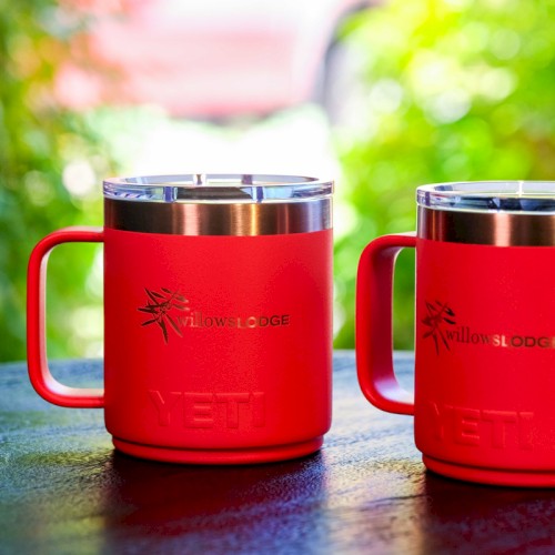 Two red Yeti mugs with handles are placed on a table with a blurry green background.