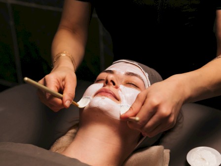 A spa facial treatment: a masked client relaxing while a practitioner applies masks with brushes at a dim, serene salon.