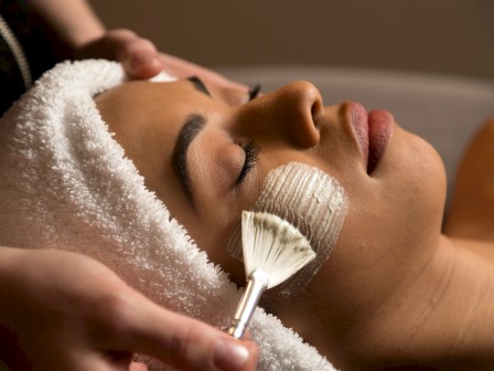 A woman receives a facial treatment, with a towel wrapped around her head, while an esthetician applies something with a fan brush.