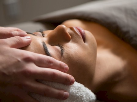 A woman receives a relaxing facial massage while lying down with eyes closed, covered by a towel, in a calm spa environment.