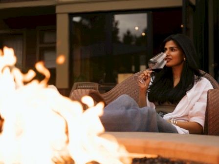 A woman with long dark hair drinks red wine next to a cozy firepit outdoors at dusk.