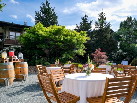 An outdoor garden setup with round tables, white cloths, floral centerpieces, wooden chairs, and barrels with drink dispensers under a sunny sky.