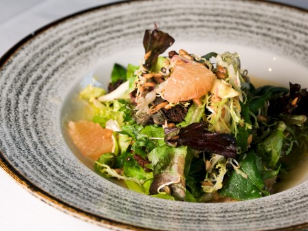 A fresh mixed greens salad with grapefruit pieces and seeds, served in a textured, round bowl.