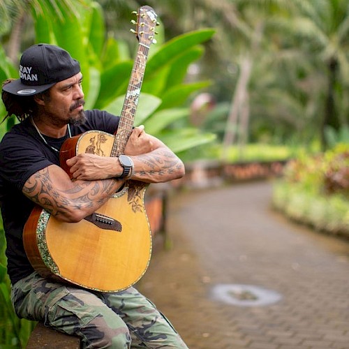 A man with a sideways hat sitting down and hugging a guitar