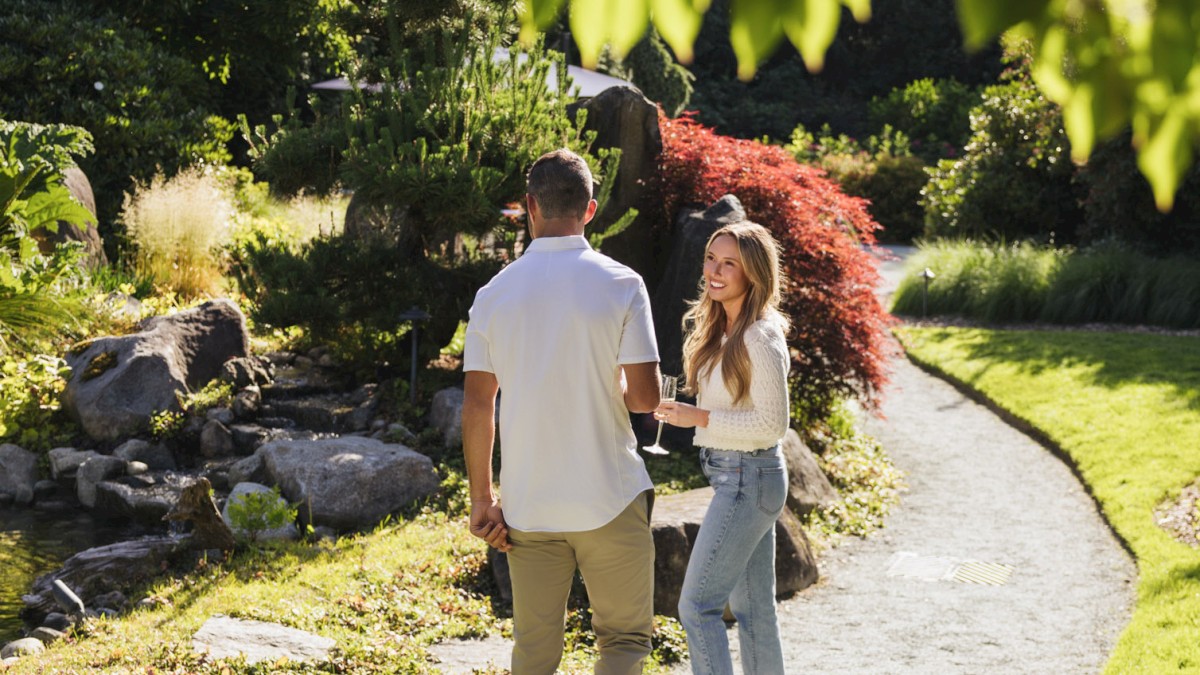A couple is enjoying a sunny moment in a lush, vibrant garden with trees, shrubs, and a winding path, sharing a drink and smiling.