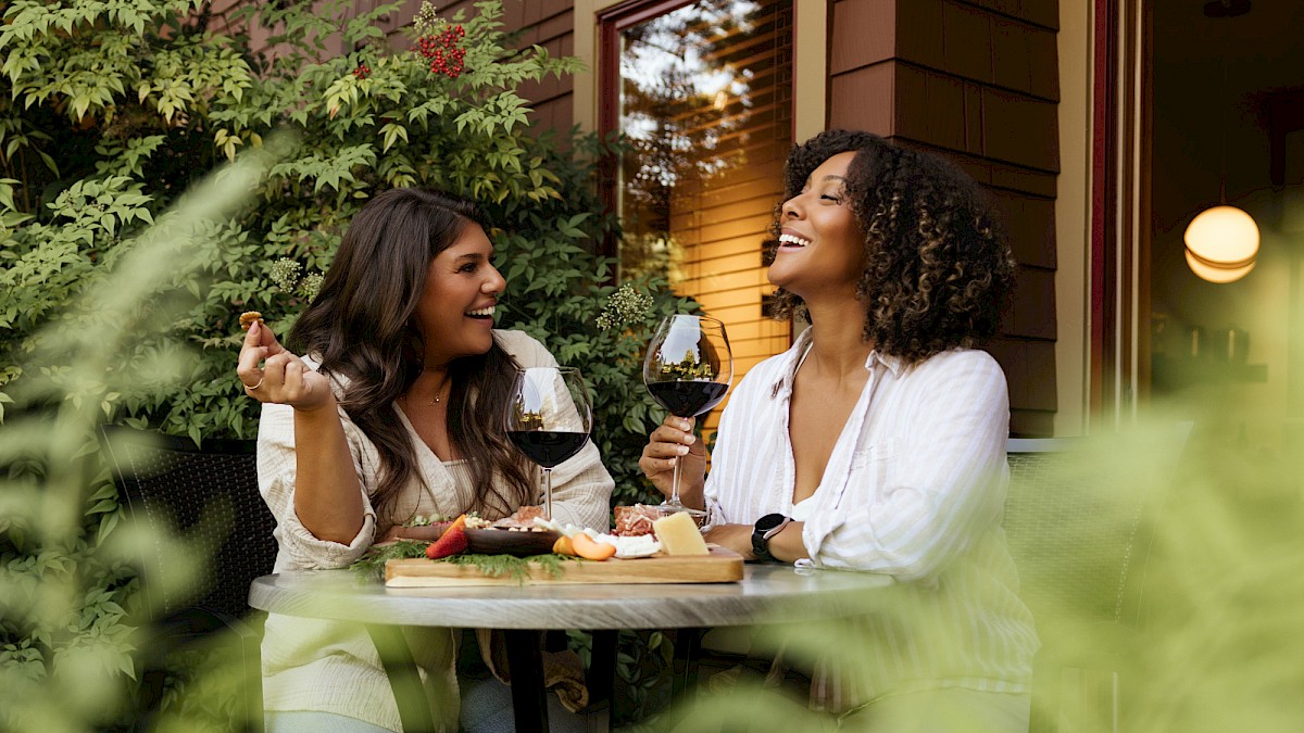 Two women are enjoying wine and cheese outdoors, sharing a joyful moment at a round table in a lush garden setting.