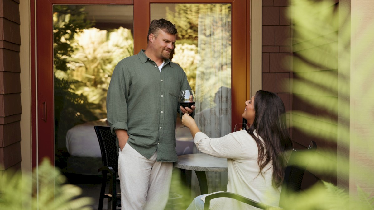 A woman offers a man a glass of red wine on a patio, creating a warm, intimate moment between them.