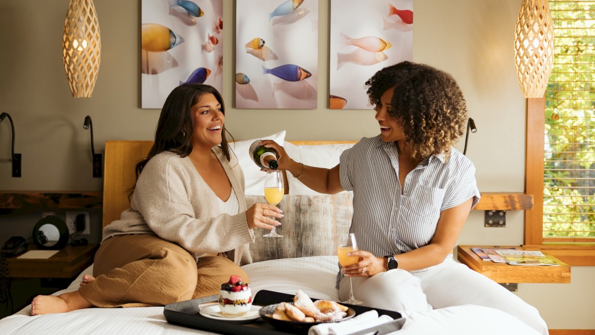 Two friends toast and share a cute breakfast on a cozy hotel bed, with coffee and donuts on a tray and tropical fish art in the background.