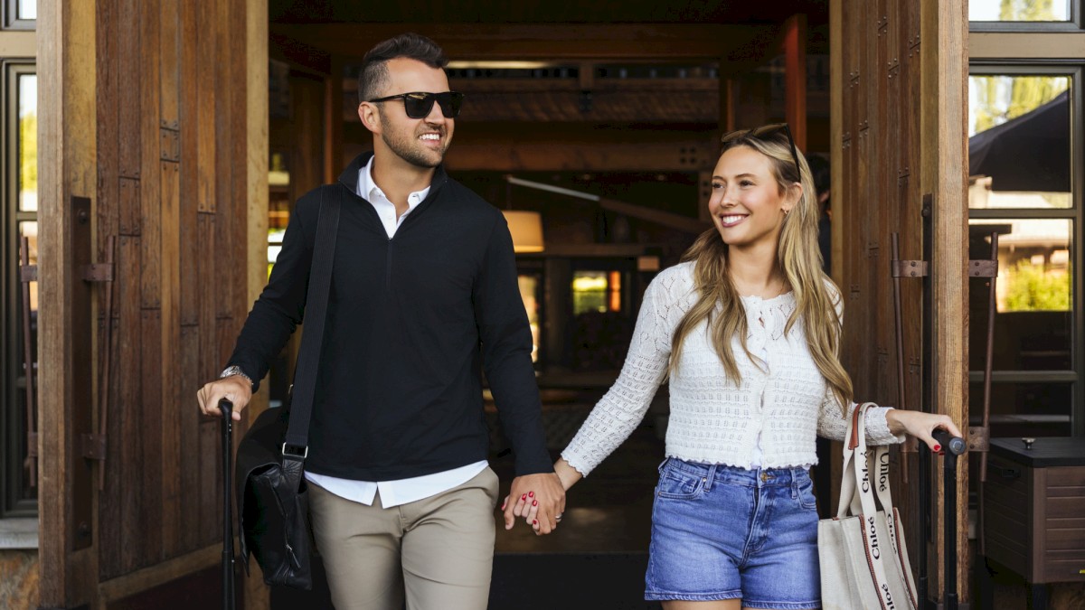 Two people walk hand-in-hand out of a doorway, smiling; man in black polo and beige pants, woman in white top and denim shorts, both carrying bags.