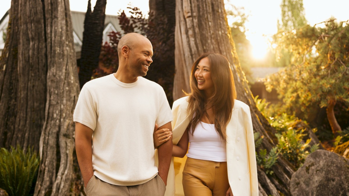 A couple walks arm-in-arm through a sunny park, smiling at each other among tall trees and warm, natural light.