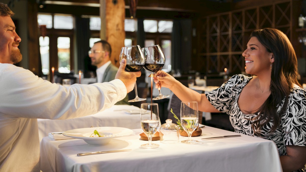 A man and woman toast with glasses of red wine in a cozy, well-lit restaurant setting.