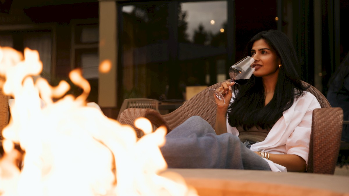 A woman lounges by a cozy fire pit, sipping wine in a white top, as flames glow warmly in the evening patio setting.