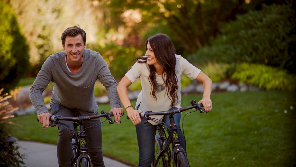 A man and woman ride bicycles on a path through a garden, smiling at each other. The setting appears sunny and lush with greenery.
