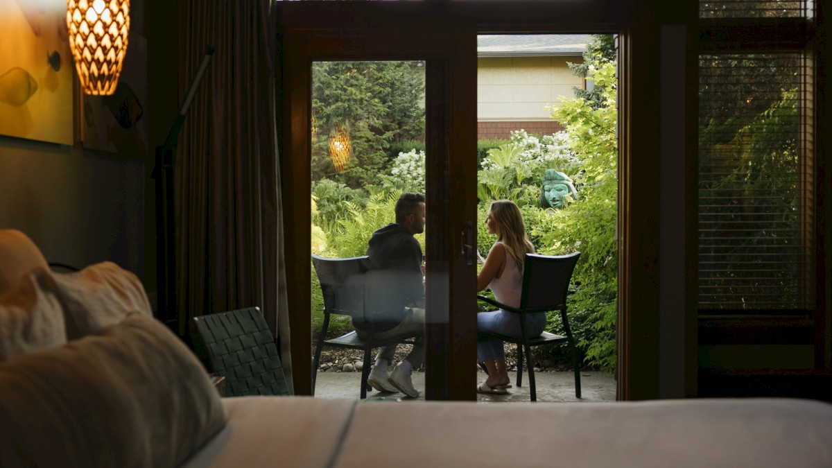 A couple is sitting outside on a patio, engaged in conversation, surrounded by lush greenery, photographed from inside a house through glass doors.