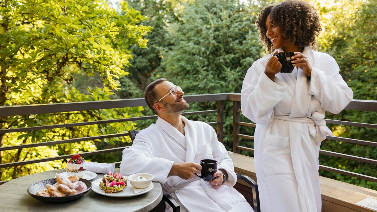 Two people in white robes enjoy coffee and desserts on a balcony surrounded by greenery, sharing a joyful moment.