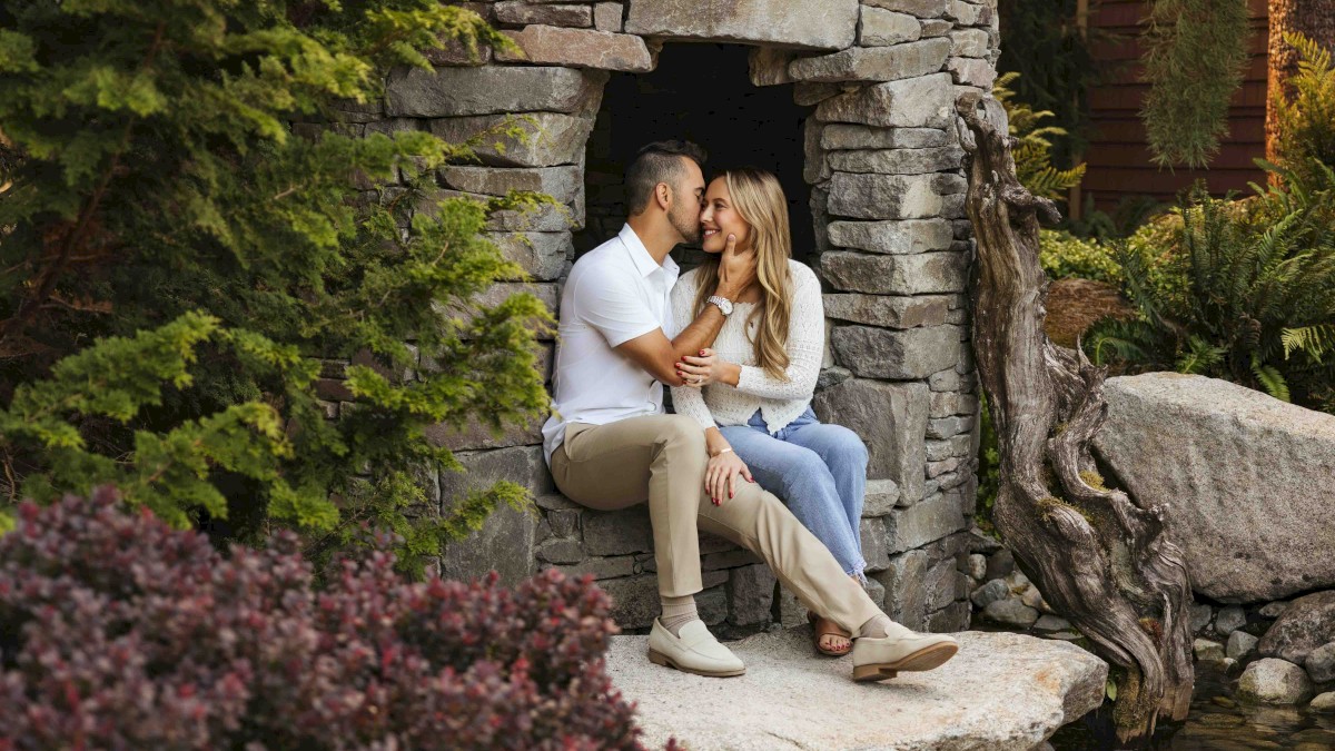 A couple sits in a stone doorway, sharing an intimate moment, surrounded by lush greenery and rocks by the water's edge.
