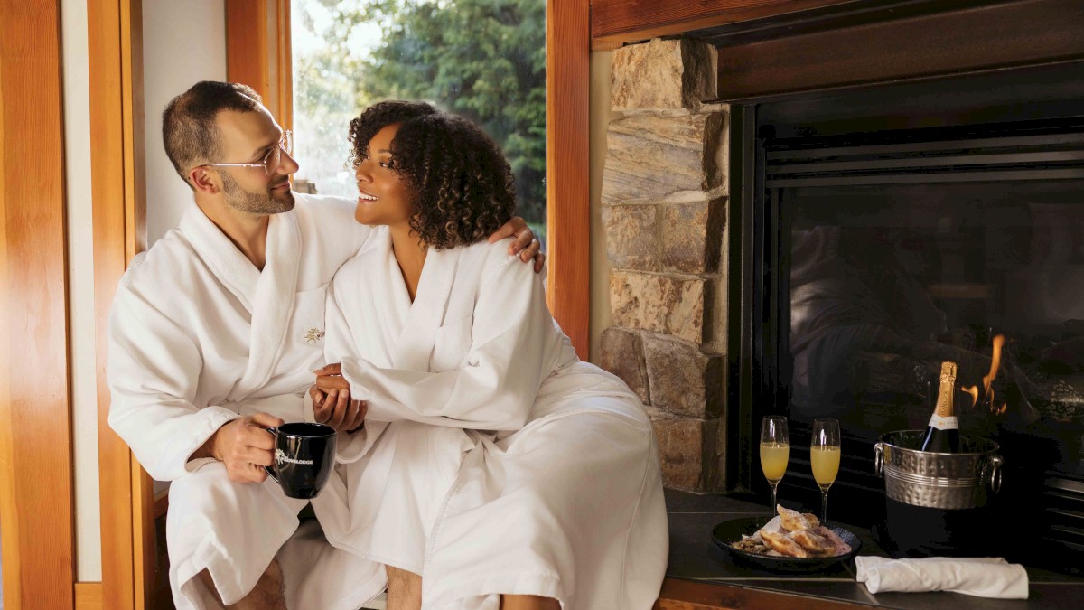 A couple in white robes sits by a fireplace, enjoying drinks and snacks. They appear relaxed and content in a cozy setting.