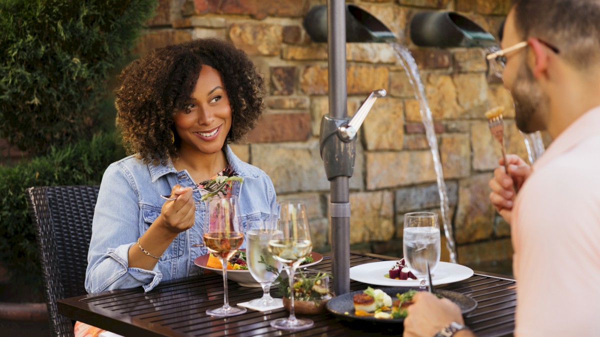 A man and woman enjoy a romantic outdoor dinner with drinks and desserts, sharing a smile beneath a water fountain feature.