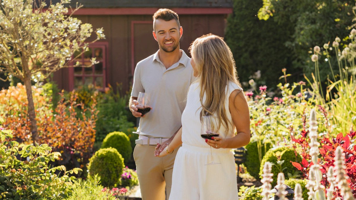 A man and woman enjoy wine in a sunny, lush garden with colorful flowers and greenery on a warm afternoon.