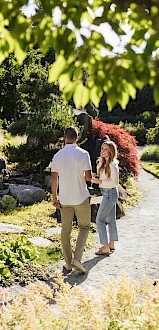 A couple is walking along a garden path surrounded by lush greenery and colorful plants on a sunny day.