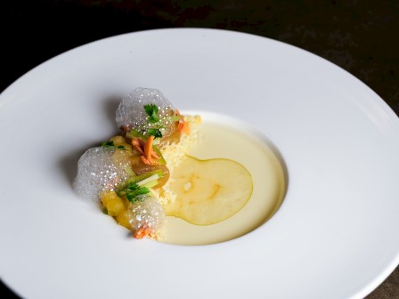 A fine dining plate with a small cluster of delicate, thin crisps and herbs arranged around a light sauce pool in the center of a white dish.