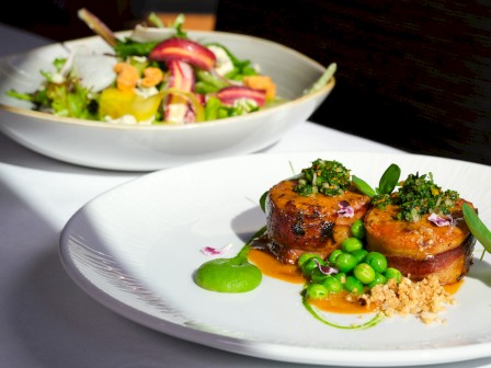 A gourmet dish featuring two seared scallops topped with herbs, served with green peas, sauce, and garnishes on a white plate.