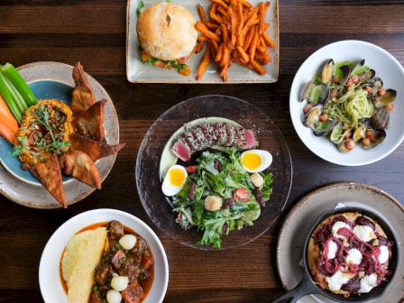 A variety of dishes including salad, burger, sweet potato fries, curry, pizza, and hummus platter on a wooden table.