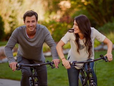A man and woman are happily riding bicycles side by side on a path, surrounded by lush greenery in a park setting.