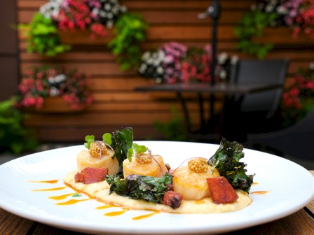 A gourmet dish with scallops, greens, bacon, and sauce, presented on a white plate outdoors with a wooden table and floral background.