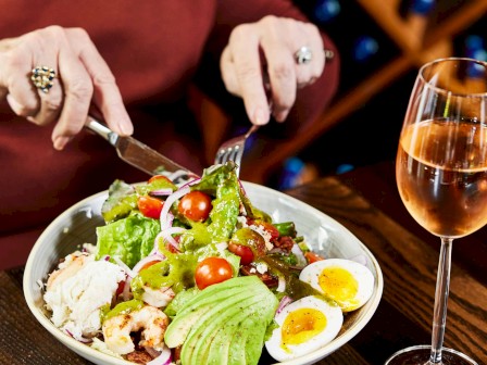 A person is enjoying a salad with avocado, eggs, and tomatoes, alongside a glass of wine on a wooden table.