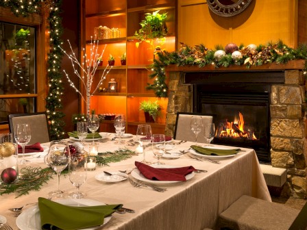 A cozy dining room decorated for Christmas with a table set, greenery, ornaments, and a lit fireplace.