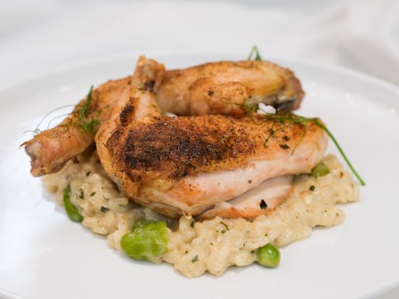 Grilled chicken pieces are served on a bed of creamy risotto with green vegetables on a white plate.