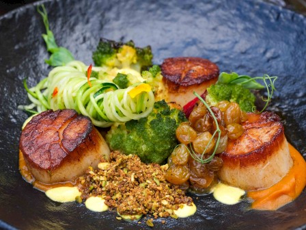 Seared scallops with spiralized vegetables, broccoli, raisins, nuts, and a creamy sauce, artfully arranged on a dark plate.