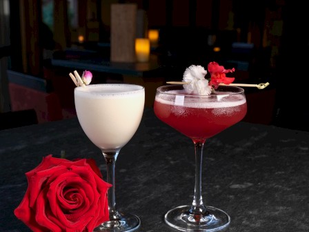 Two cocktails on a dark table with a red rose, one white and one red, garnished with flowers; romantic bar vibe.