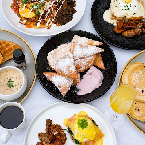 A spread of breakfast foods including waffles, tacos, pastries, pancakes, a savory dish, and beverages is shown on a white table.
