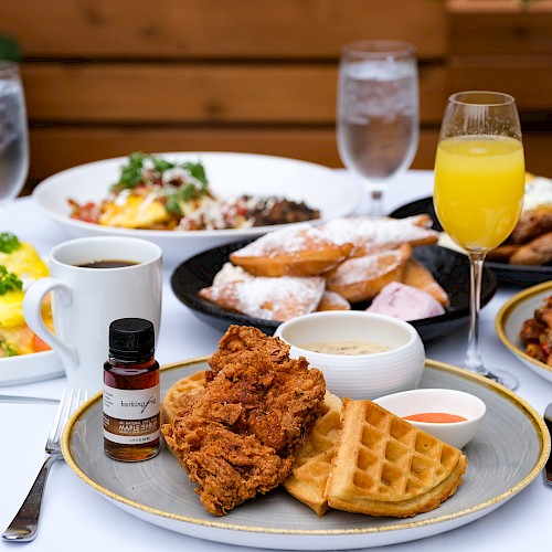 A table set with brunch items: fried chicken, waffles, beignets, eggs, bacon, coffee, orange juice, and a small syrup bottle.
