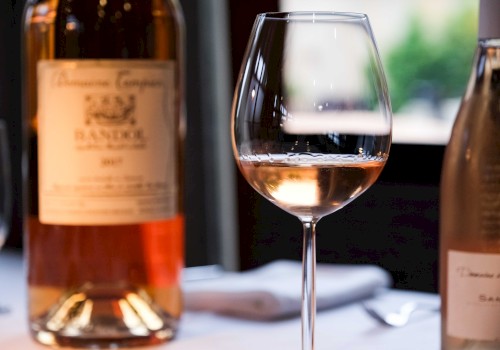 A glass of rosé wine is on a table next to a bottle with a blurred background view through a window in soft lighting.