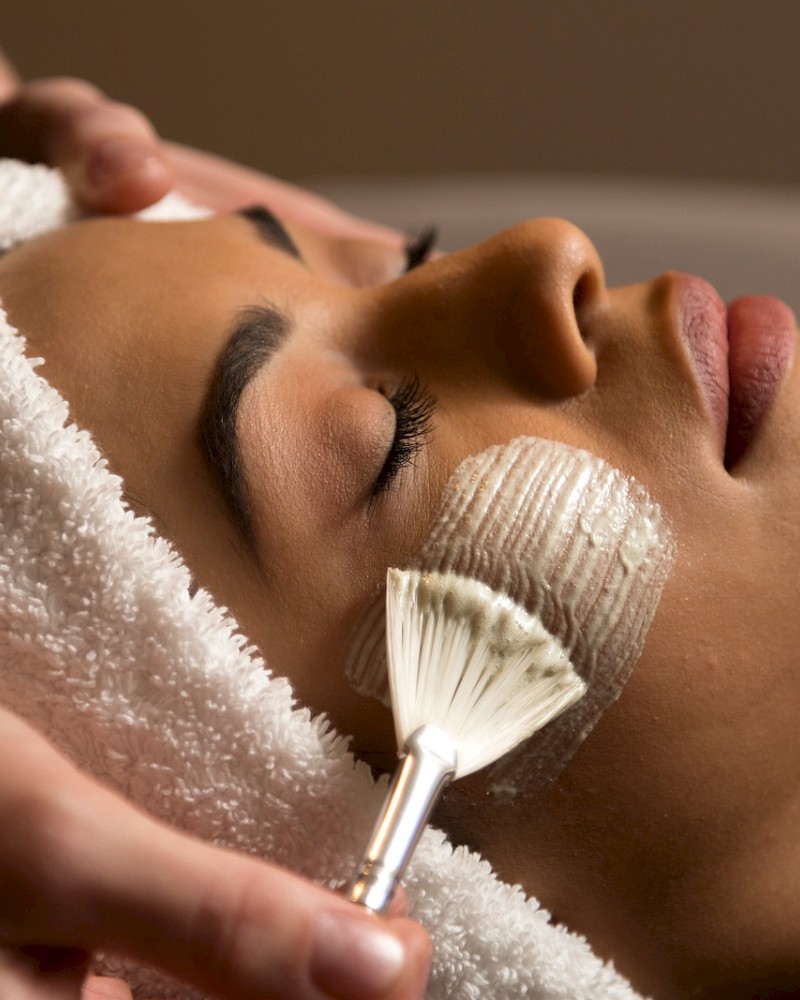 A person is receiving a facial treatment, with a towel on their head and a brush applying a mask to their cheek.