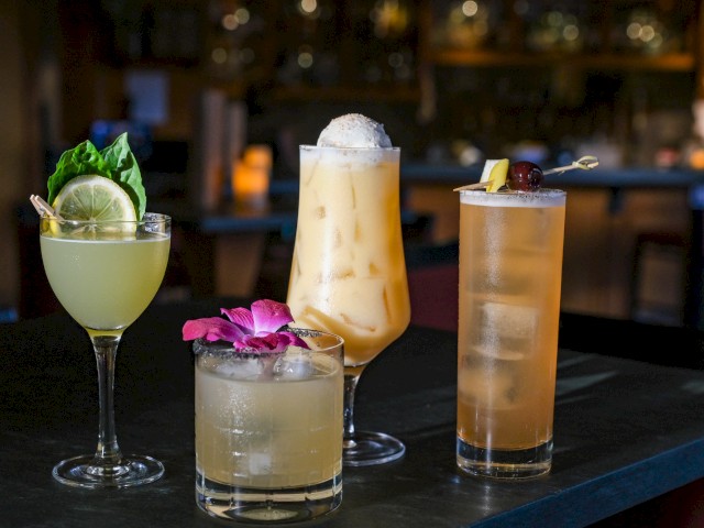 Four cocktails on a bar: a lime-basil garnish, a pink flower garnish, creamy with foam, and a tall drink with a cherry garnish.