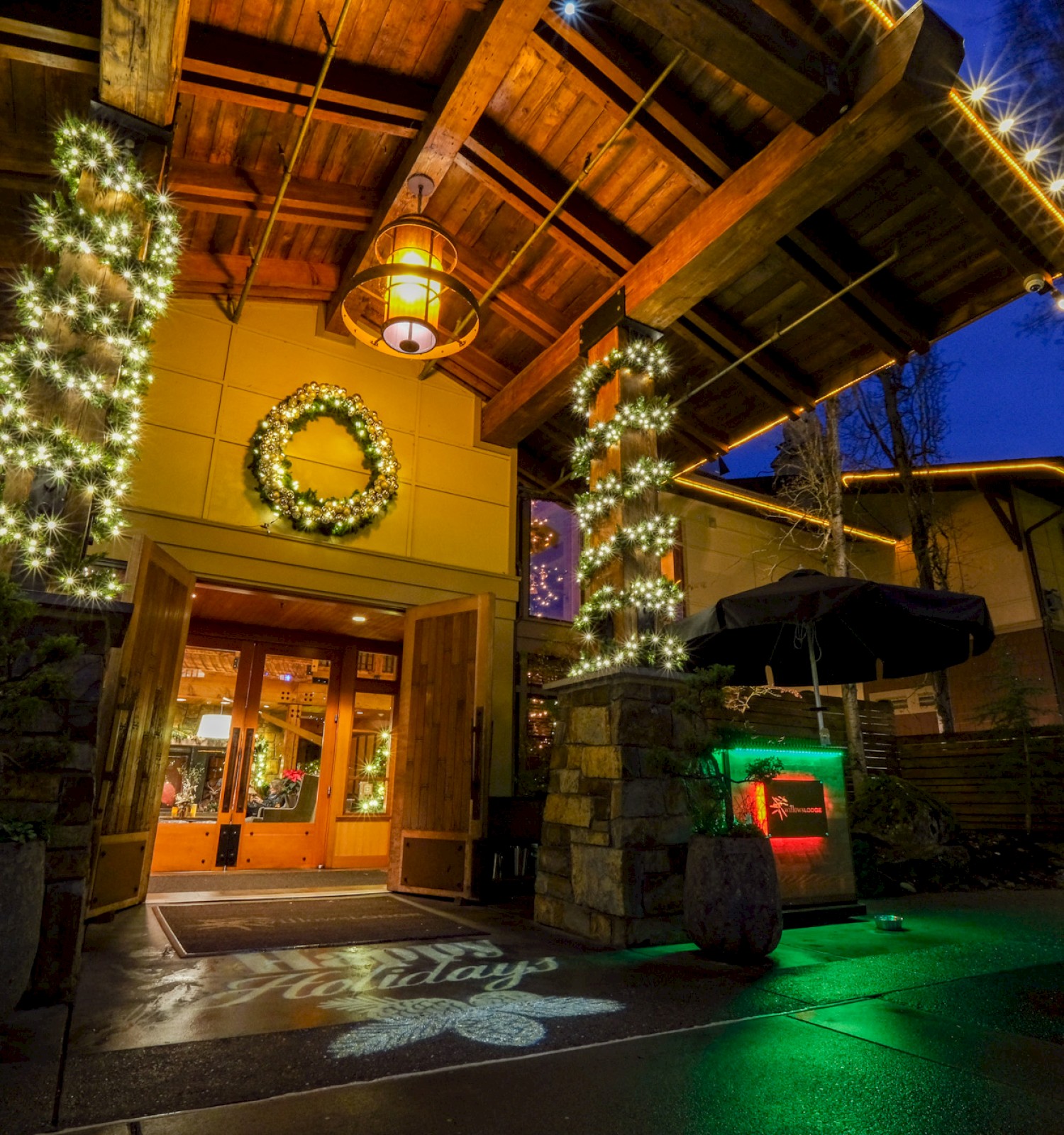 A warmly lit building decorated with Christmas wreaths, lights, and a tree, suggesting a festive holiday atmosphere at night.