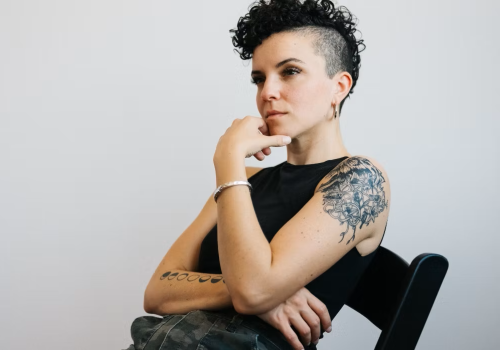 A person with curly hair and tattoos sits on a chair, contemplating.