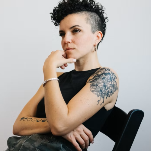 A person with curly hair and tattoos sits on a chair, contemplating.