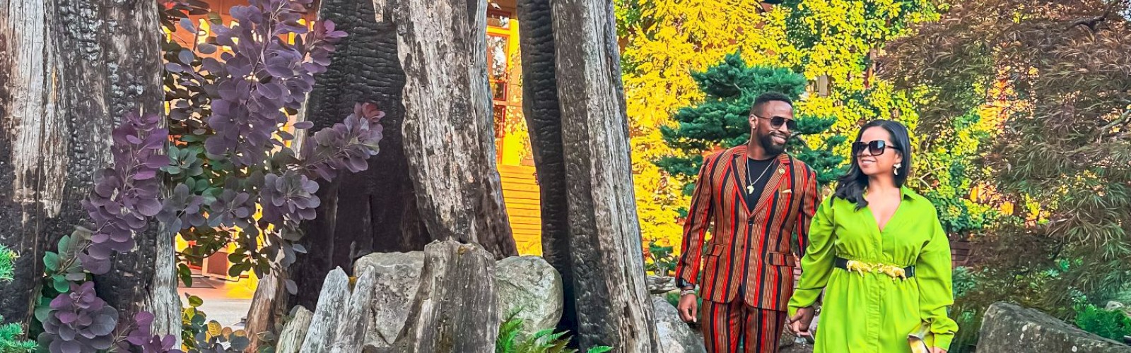 A couple walks through a vibrant garden with tall trees and yellow flowers lining a pathway, creating a picturesque scene.