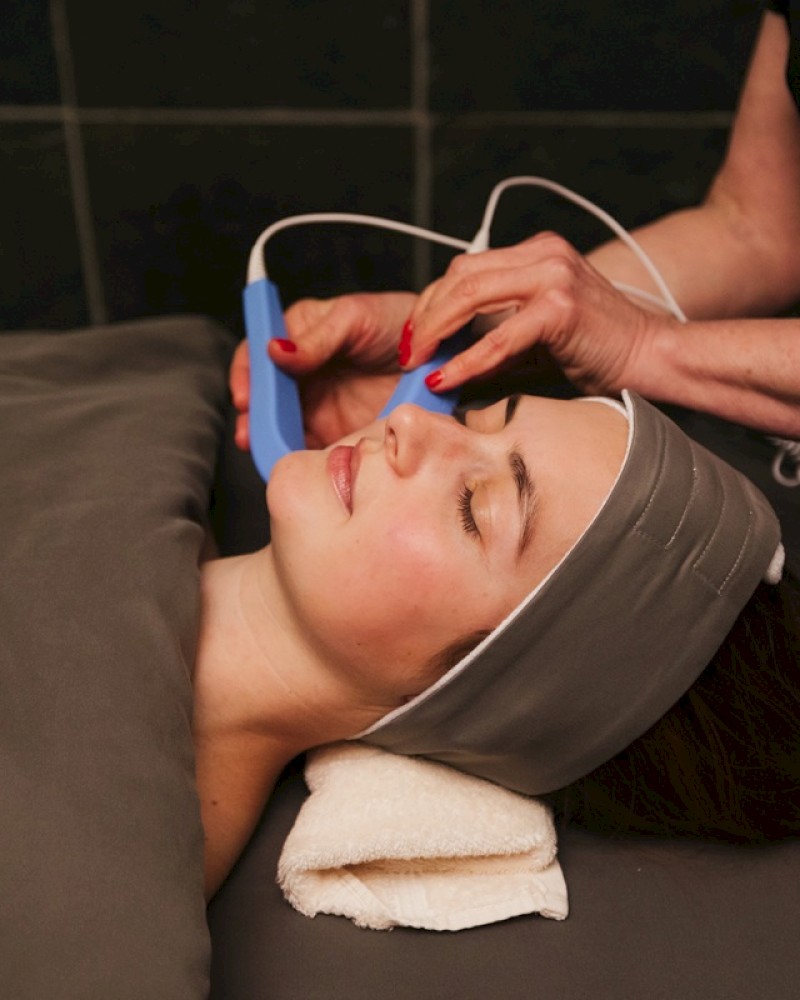 A person is receiving a facial treatment with a handheld device in a spa-like setting, lying down and covered with a blanket.