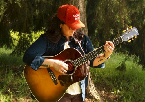 A person in a red cap plays an acoustic guitar while standing in a sunlit forest setting, surrounded by green grass and trees.