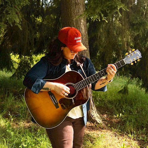 A person in a red cap plays an acoustic guitar while standing in a sunlit forest setting, surrounded by green grass and trees.