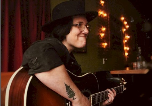 A person with glasses and a hat plays an acoustic guitar in a dimly lit room with string lights and a tree tattoo on their arm.