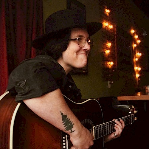 A person with glasses and a hat plays an acoustic guitar in a dimly lit room with string lights and a tree tattoo on their arm.